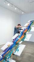 Young male artist creating colorful paint flow installation in modern white gallery space with track lighting and concrete floors.