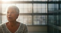 Senior woman with short gray hair and light skin tone, wearing a light sweater, gazing thoughtfully toward a window with urban cityscape visible behind her, suitable for lifestyle and corporate concepts.