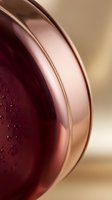 Closeup of burgundy liquid in glass sphere with water droplets and soft beige background, suitable for cosmetics and skincare product packaging design.