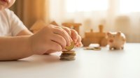 Child's hand stacking coins into a tower on white surface with wooden toys in soft sunlight, representing savings, financial education, and money management for kids.