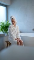 Young Asian woman in white bathrobe and towel sitting by bathtub in modern spa bathroom with fern plant enjoying relaxing self-care wellness routine.