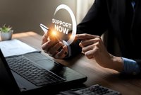 Professional businessman holding glowing support now icon above laptop offering customer service assistance in modern office workspace.