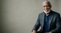 Senior African American male professional with gray beard wearing dark blazer and blue shirt, smiling confidently against neutral background. Suitable for corporate, business, leadership, and professional services marketing materials.