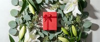 Red gift box with ribbon surrounded by white lilies and eucalyptus branches on white background for holiday celebrations and special occasions.