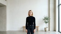 Young Caucasian woman with blonde hair wearing black long-sleeve top standing in minimalist interior with potted plant and wooden stool, suitable for corporate, lifestyle, and professional website design.