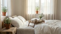 Cozy bedroom interior with white bedding, wooden breakfast tray, potted plants on windowsill, natural light filtering through curtains creating peaceful morning atmosphere.