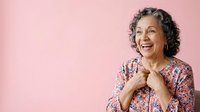 Mature woman with gray curly hair and warm smile wearing floral print shirt against pink background, expressing joy and positivity for lifestyle and wellness content.