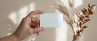 Hand holding blank white business card mockup with dried pampas grass and eucalyptus branches in warm natural light for branding design presentation.
