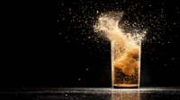 Dramatic splash of golden liquid in glass against dark background with floating particles creating dynamic motion effect and light reflection.