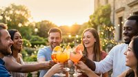 Diverse group of young adults toasting with colorful cocktails on outdoor patio during golden hour sunset celebration with friends laughing together.
