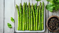 Fresh green asparagus spears on white plate with black peppercorns and basil leaves against rustic wooden background. Natural organic vegetables for healthy eating.