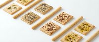 Assorted crackers with seeds and herbs arranged in diagonal rows with wooden spreaders on white background for food styling.