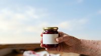 Elderly person with fair skin holding a glass jar with blank white label and dark red jam or preserve against rural landscape background for product packaging and branding.