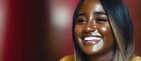 Young African American woman with long ombre hair smiling genuinely while wearing yellow sweater against blurred red background, showing bright white teeth.