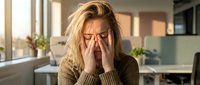 Stressed blonde businesswoman with glasses covering face with hands experiencing headache or fatigue in modern office workspace during workday.