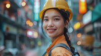 Young Asian female construction engineer wearing yellow hard hat and safety vest smiles confidently on urban street with bokeh lights in background.