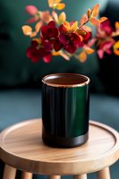 Elegant dark green ceramic mug with copper rim on wooden round table, red and orange orchid flowers in soft focus background creating cozy atmosphere.