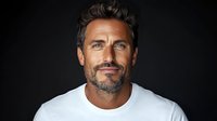 Confident mature Caucasian male with salt and pepper beard wearing white t-shirt against dark background, smiling at camera. Professional headshot style portrait.