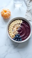 Acai bowl topped with sliced banana, blueberries, and almonds, served with fresh peeled mandarin and water glass on marble surface for healthy breakfast or smoothie bowl content.