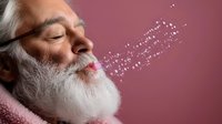 Senior Caucasian man with white beard blowing glitter dust against pink background, creating magical sparkle effect in studio lighting. Conceptual aging gracefully.