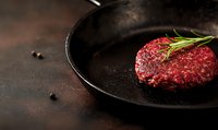 Raw beef burger patty with fresh rosemary sprig on black plate against dark rustic background with peppercorns, dramatic food photography for restaurant menu.