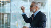 Mature Caucasian businessman with gray hair and glasses writing strategy notes on glass wall in modern office, focused on business planning and analysis.