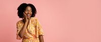 Surprised African American woman with curly hair in yellow floral dress touching face with hand against pink background for lifestyle marketing.