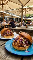 Two gourmet pulled pork sandwiches with purple cabbage slaw on blue plates at outdoor restaurant patio with umbrellas and diners in background.