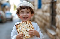 Smiling young boy in traditional white clothing holding matzo bread during Jewish Passover celebration on urban street.