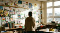 Teacher in brown sweater viewing colorful student artwork display wall in bright classroom with natural light from large windows.