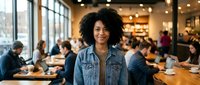 Young African American woman with curly hair smiling in busy modern cafe restaurant. Professional portrait for business, hospitality industry marketing.
