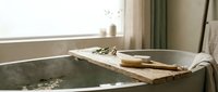 Relaxing spa bathroom scene with wooden bath caddy holding natural brush and towels across modern bathtub by bright window for wellness and self-care moments.