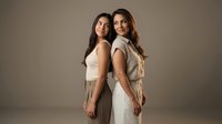 Two young women standing back to back in elegant neutral outfits against beige background for fashion and lifestyle photography.