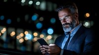 Mature bearded businessman in suit checking smartphone at night, illuminated by screen glow against blurred city lights background. Professional corporate portrait.