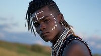 Young African male model with tribal face paint, braided hair, and traditional beaded necklace poses against natural outdoor setting during golden hour lighting.
