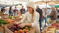 Young woman in beige knit hat shopping for fresh organic vegetables at outdoor farmers market with wooden crates full of colorful produce and shoppers.
