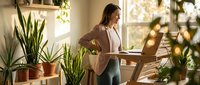 Young woman working on laptop in bright home office surrounded by green plants and natural sunlight creating productive workspace atmosphere.