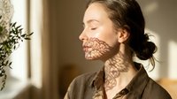Young Caucasian woman with closed eyes enjoying warm sunlight streaming through window creating leaf shadow patterns on her face in cozy home interior.