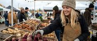Young blonde woman in beige knit hat and brown apron shopping for fresh produce at outdoor farmers market with wooden crates of vegetables and fruits.