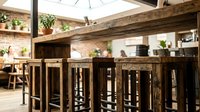 Rustic wooden bar counter with tall stools in modern loft interior featuring exposed beams, skylights, and potted plants for hospitality design.