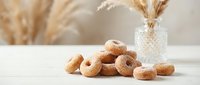 Fresh glazed donuts arranged on white surface with pampas grass decoration in crystal vase creating warm cozy bakery atmosphere for food photography.