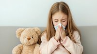Young woman with long brown hair sneezing into tissue while sitting on gray couch next to plush teddy bear, wearing pink sweater, showing cold and flu symptoms.