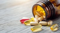 Prescription medication pills and supplements spilling from brown glass bottle onto wooden surface, colorful capsules in red and yellow tones with dramatic lighting.