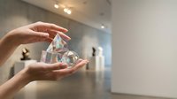 Female hands holding transparent crystal pyramid in modern gallery space with soft lighting and blurred artwork background for meditation and wellness concepts.