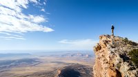 Lone hiker stands on dramatic cliff edge overlooking vast desert canyon landscape with red rocks and distant mountains under blue sky with white clouds.