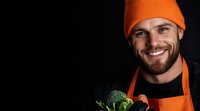 Cheerful male chef in orange beanie and apron holding fresh broccoli against dark background. Professional food service worker with beard showing organic produce.