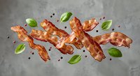 Crispy bacon strips with fresh basil leaves and black peppercorns scattered on textured surface for culinary and food photography projects.