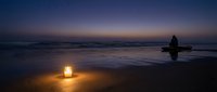 Solitary figure sits by glowing lantern on twilight beach with purple sky and gentle waves reflecting warm light on wet sand creating peaceful meditation scene.