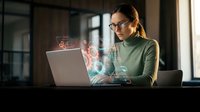 Young professional woman with glasses working on laptop with holographic digital interface elements in modern office space for technology and business concepts.