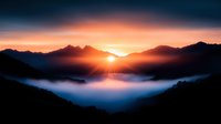 Dramatic mountain sunrise over misty valley with vibrant orange sky and sunburst breaking through peaks, creating ethereal morning atmosphere in wilderness landscape.